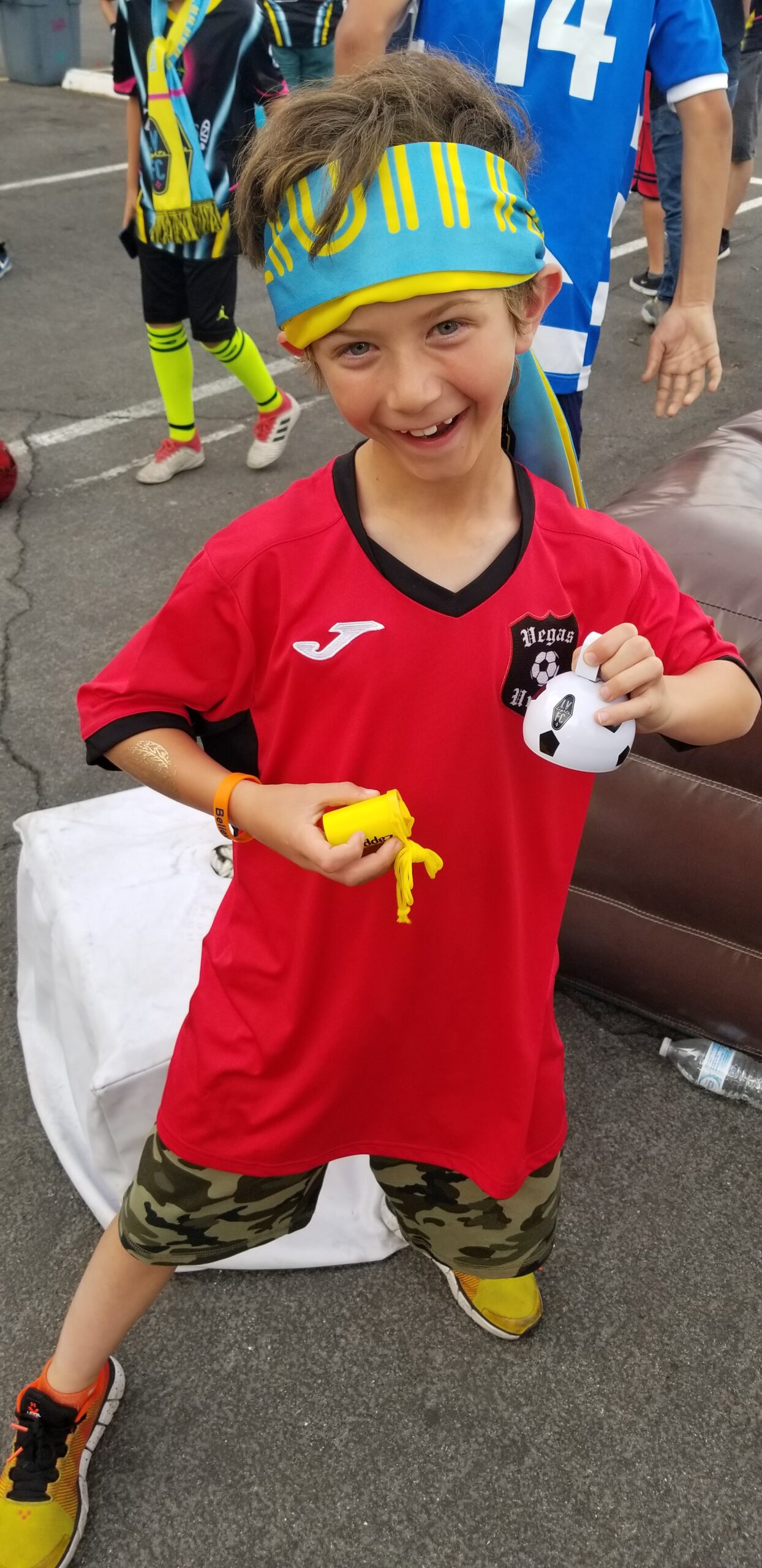 Child wearing a red soccer jersey and camo shorts, holding a small soccer ball, smiling and posing.
